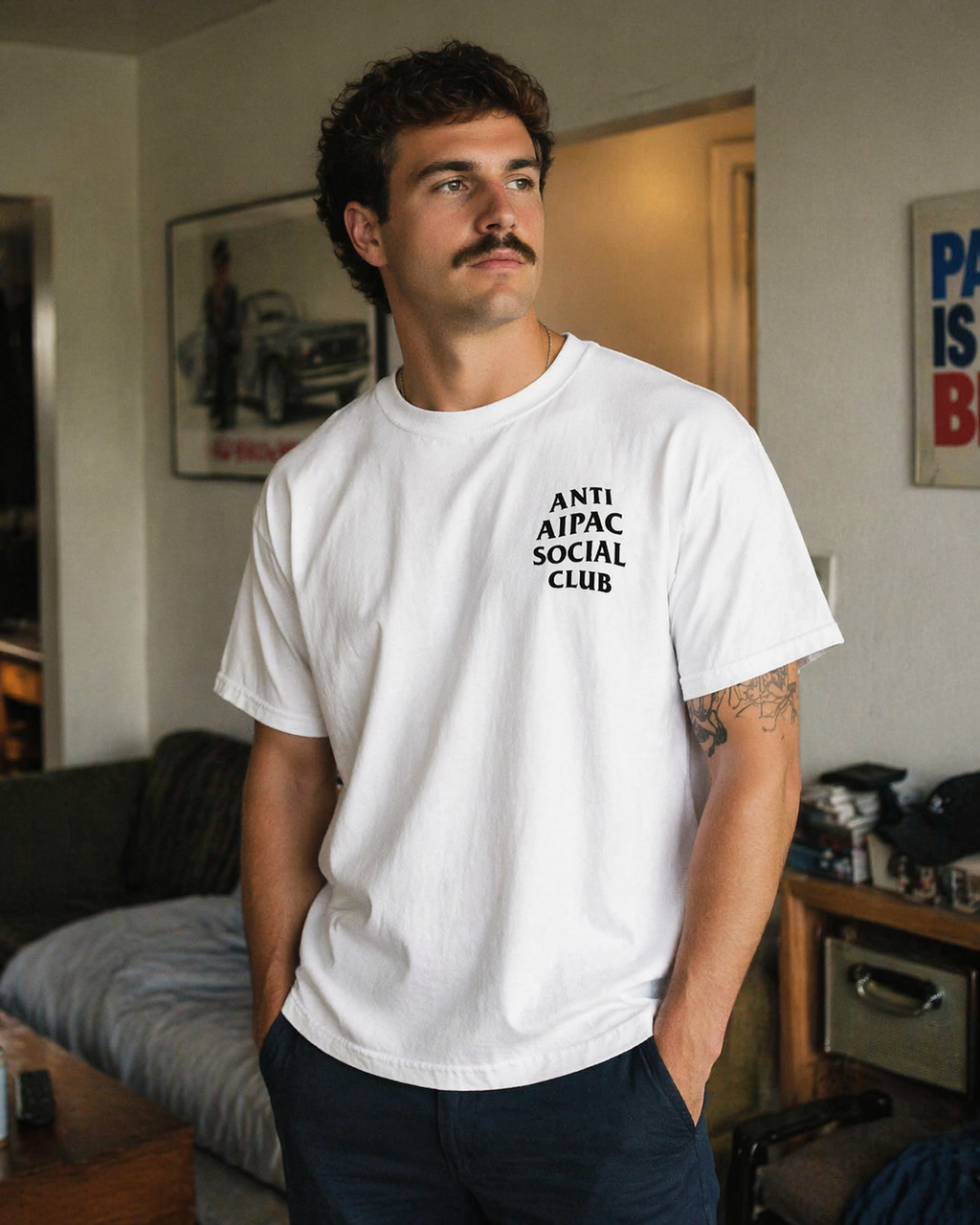 Man wearing a white t-shirt with 'ANTI AIPAC SOCIAL CLUB' text in a casual indoor setting.